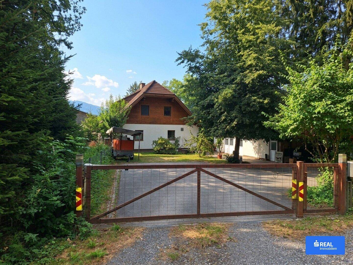 Zufahrt zum Anwesen mit einem großen Tor, umgeben von Bäumen und viel Grün.