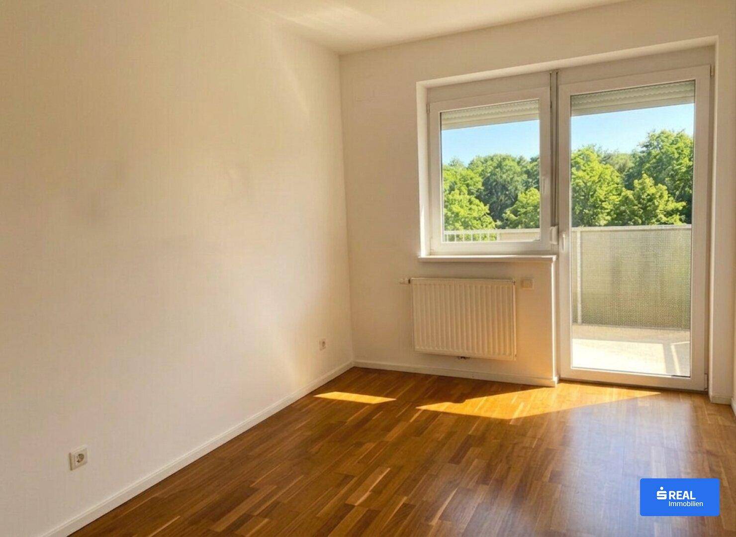 Helles Schlafzimmer mit Holzboden und Zugang zu einem Balkon, umgeben von Grün.
