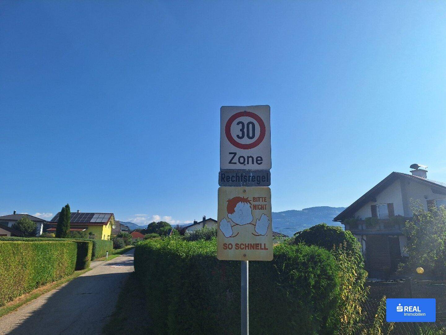 Verkehrsberuhigte Straße mit 30er-Zone-Schild und Häusern im Hintergrund, umgeben von Hecken.