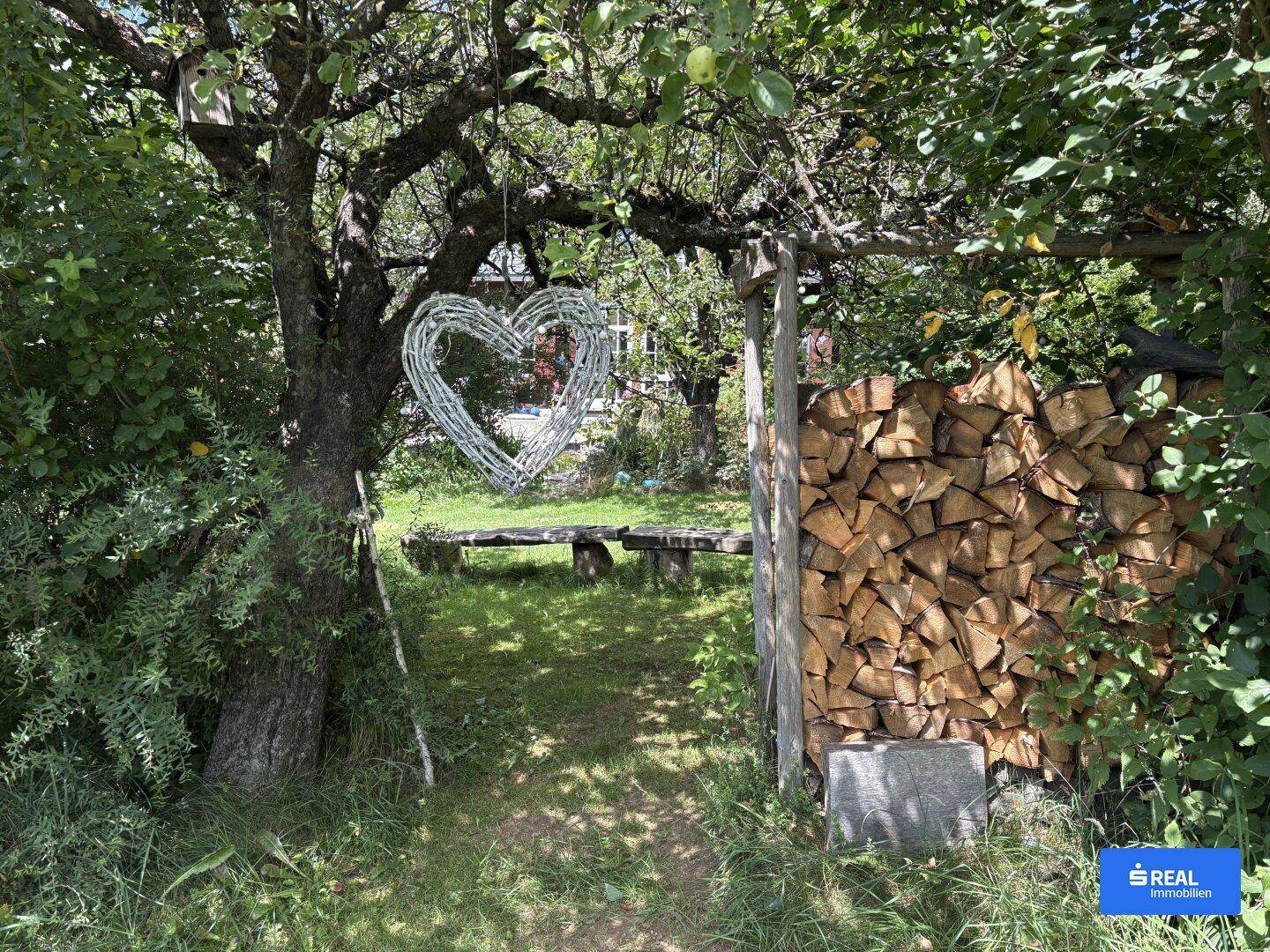 Naturbelassener Gartenbereich mit Holzstapel, Sitzgelegenheit unter Bäumen und Herzdekoration.
