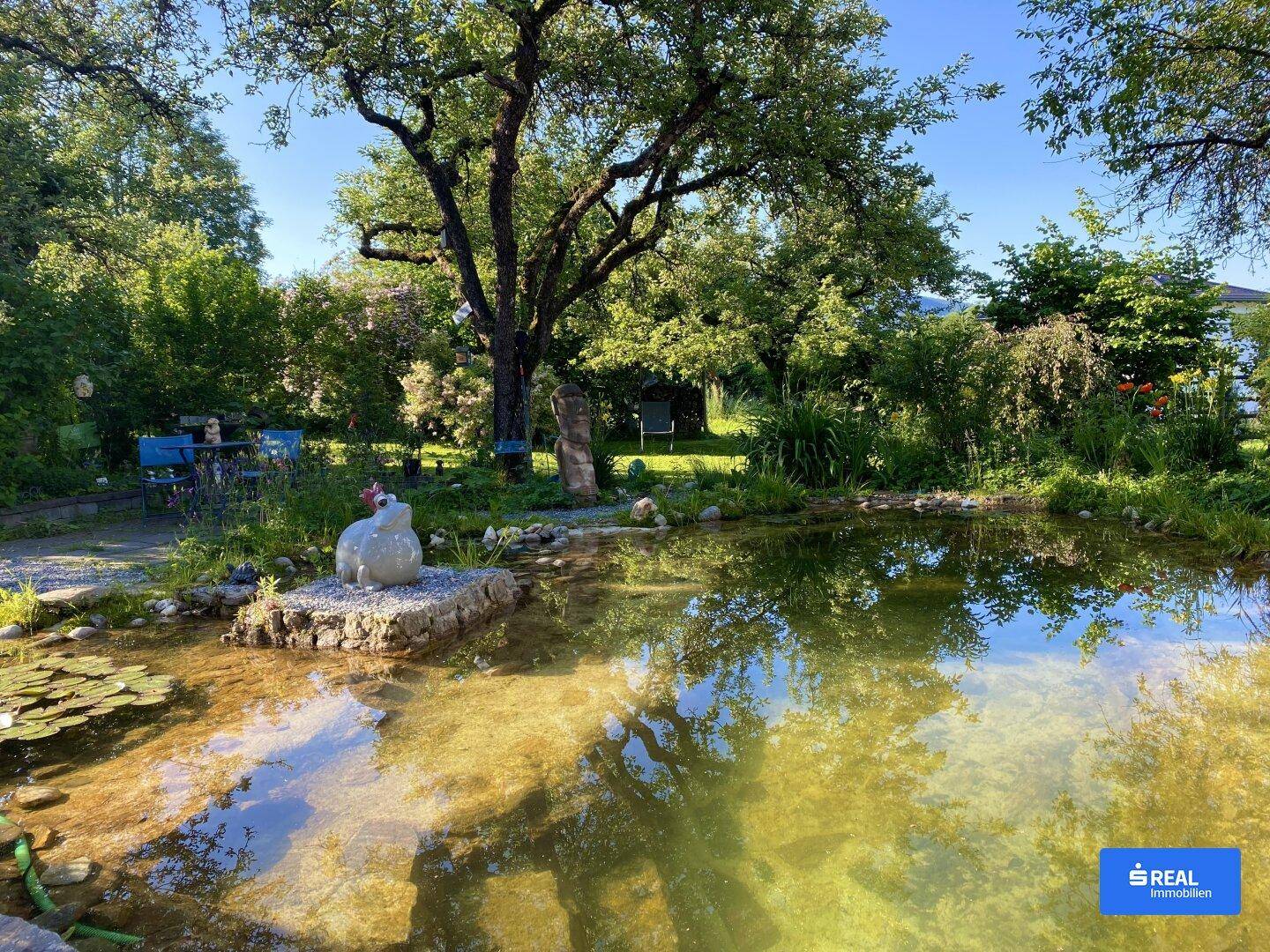 Großer Garten mit Teich, üppiger Vegetation und Sitzgelegenheiten unter schattenspendenden Bäumen.