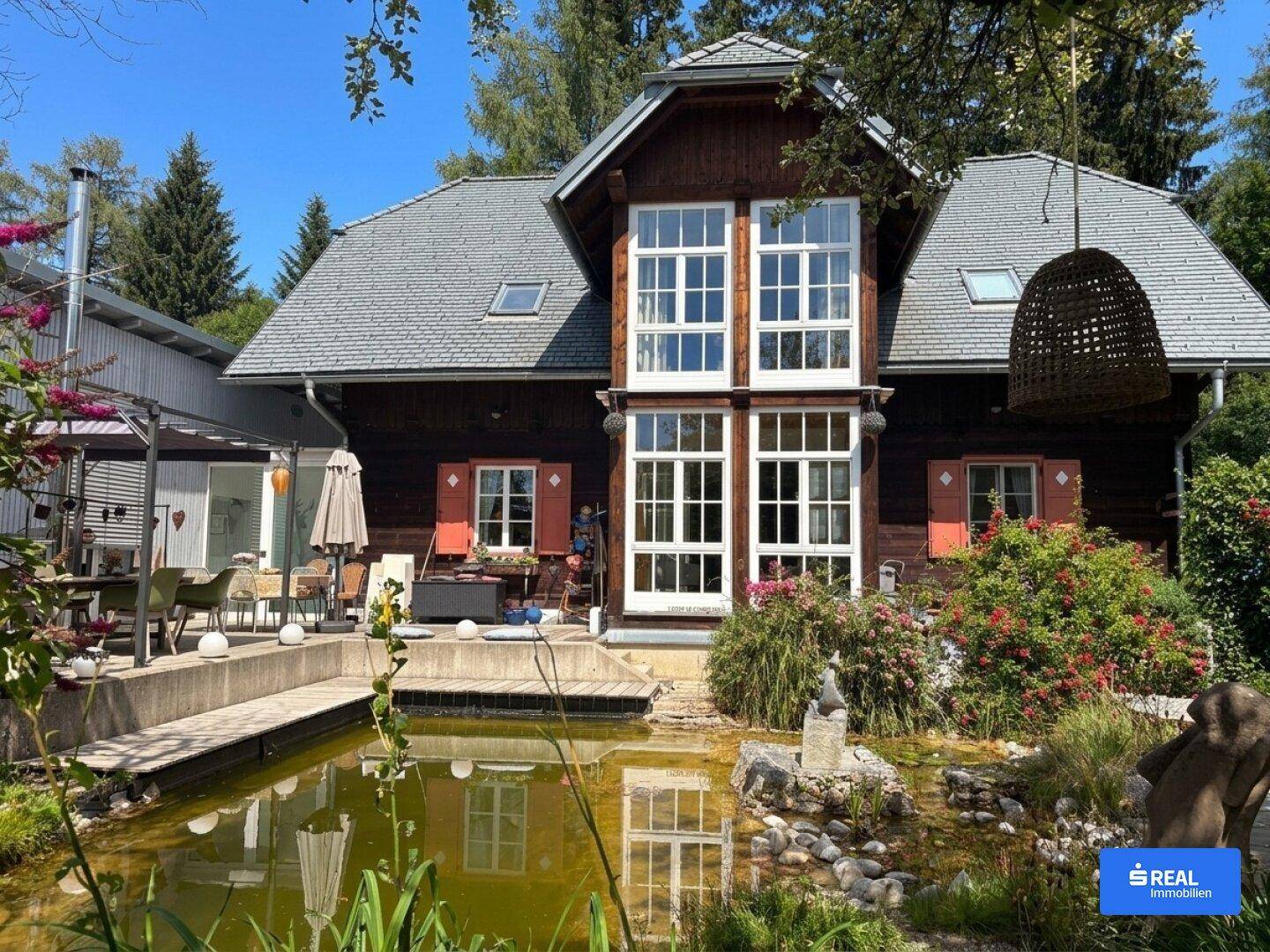 Idyllisches Holzhaus mit großem Teich und üppiger Bepflanzung im Gartenbereich.