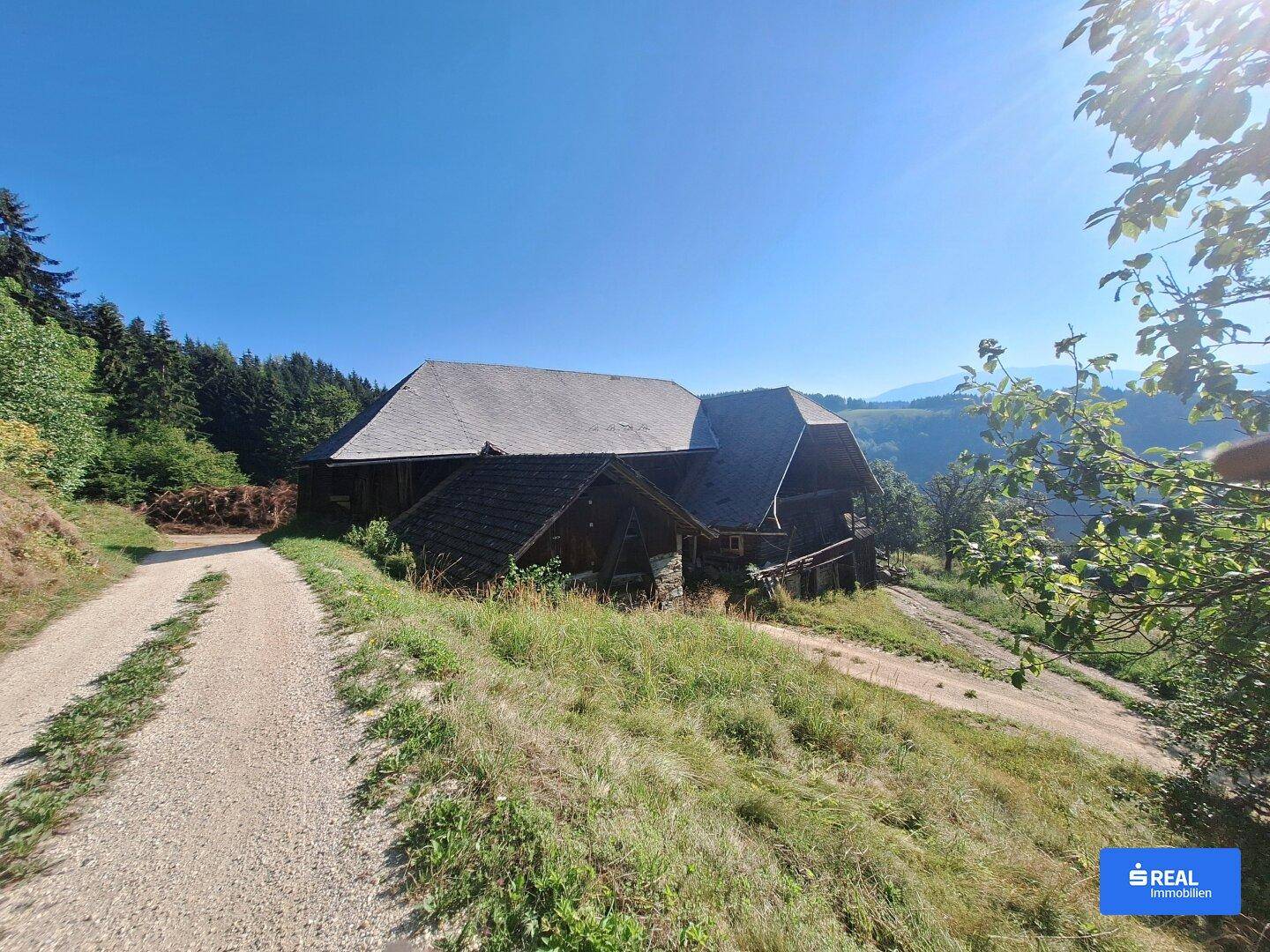 Altes, rustikales Holzgebäude auf einem Hügel mit Schotterweg und weitem Blick in die Landschaft.