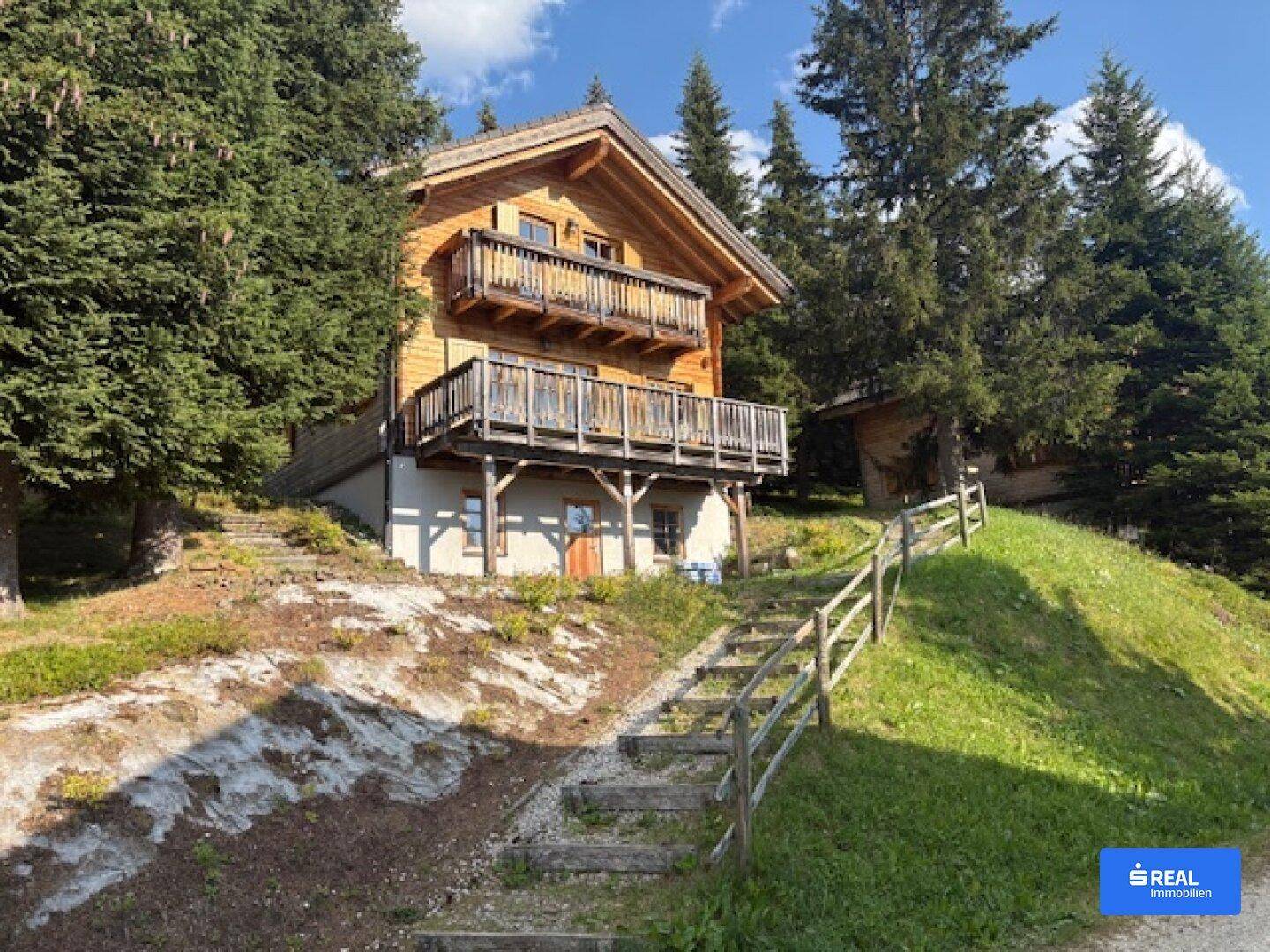 Traditionelles Holzchalet auf einem Hanggrundstück, umgeben von Bäumen, mit einem rustikalen Steintreppenaufgang.
