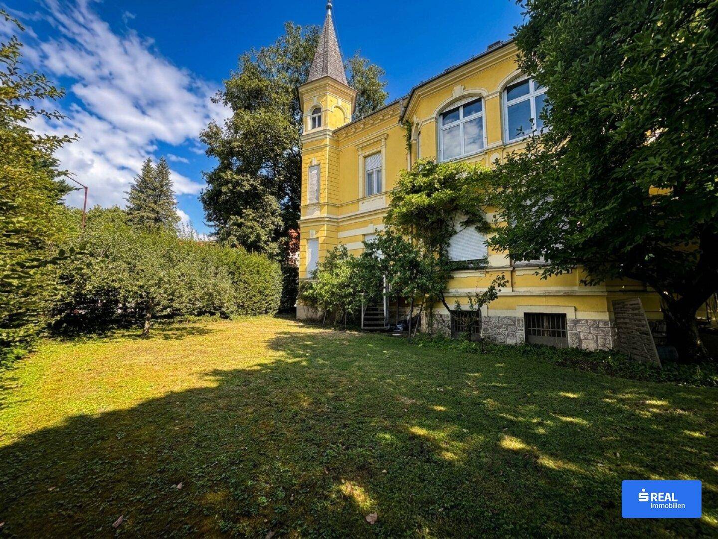 Großer Gartenbereich mit gepflegtem Rasen und altem Baumbestand neben dem historischen Gebäude mit Turm.