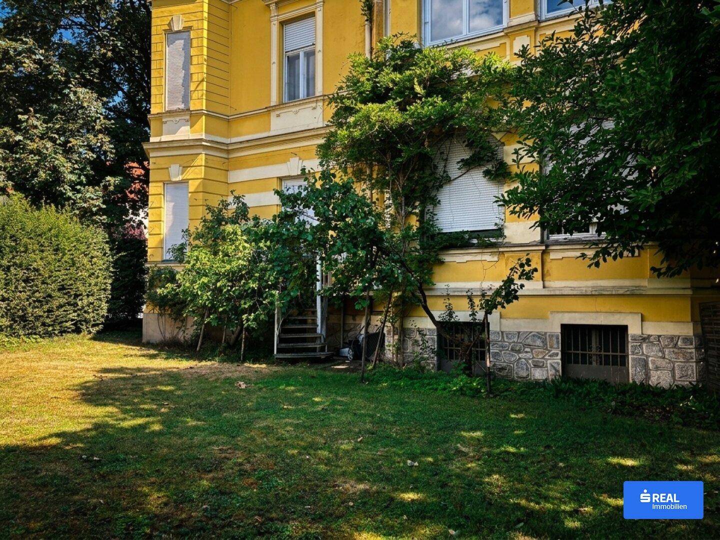 Detailansicht der Gebäudeseite mit Steinfundament, Treppe und üppiger Vegetation im Gartenbereich.