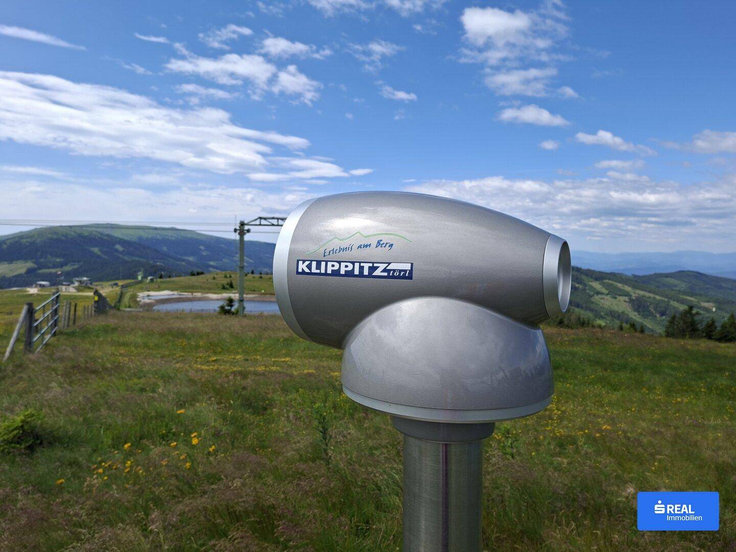 Aussichtsfernrohr auf einem Berg mit Panoramablick über die alpine Landschaft und einen See.