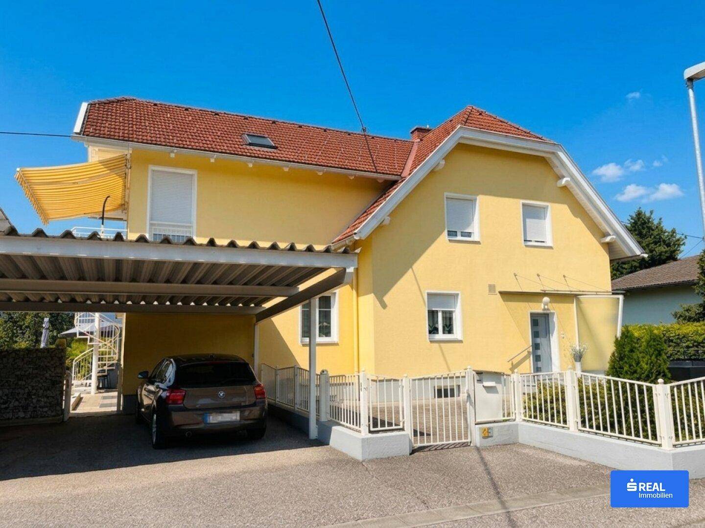 Gelbes Wohnhaus mit Carport und weißem Zaun, umgeben von einer gepflegten Einfahrt.