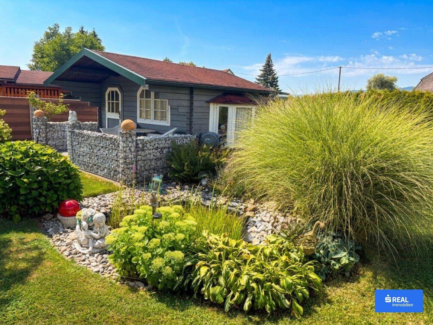 Gepflegter Garten mit einem kleinen Gartenhaus, Zierpflanzen und Gabionenmauern.