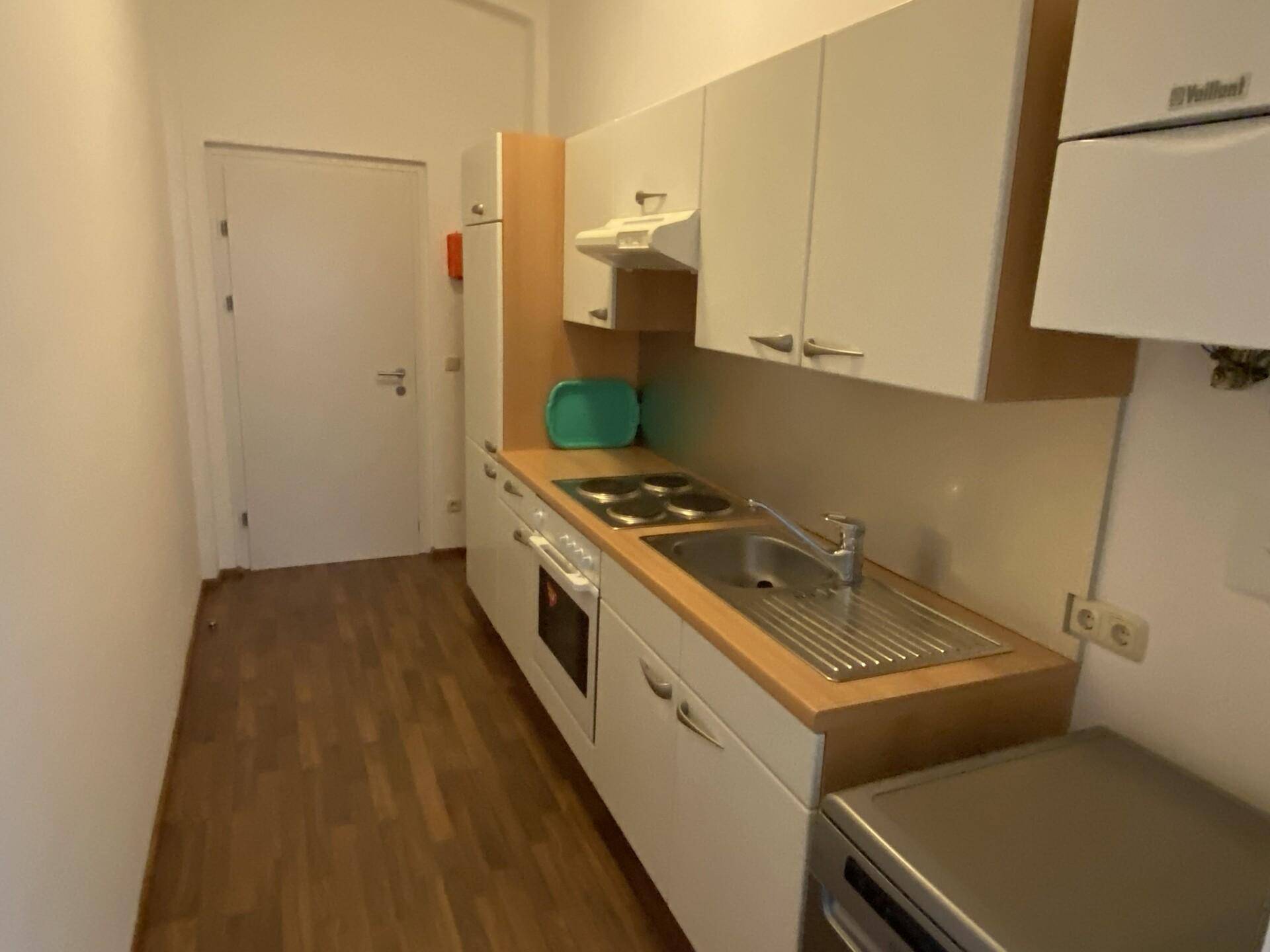 Kleine Küchenzeile mit weißen Schränken, Kochfeld, Spüle und einem Kühlschrank in einem schmalen Raum.
