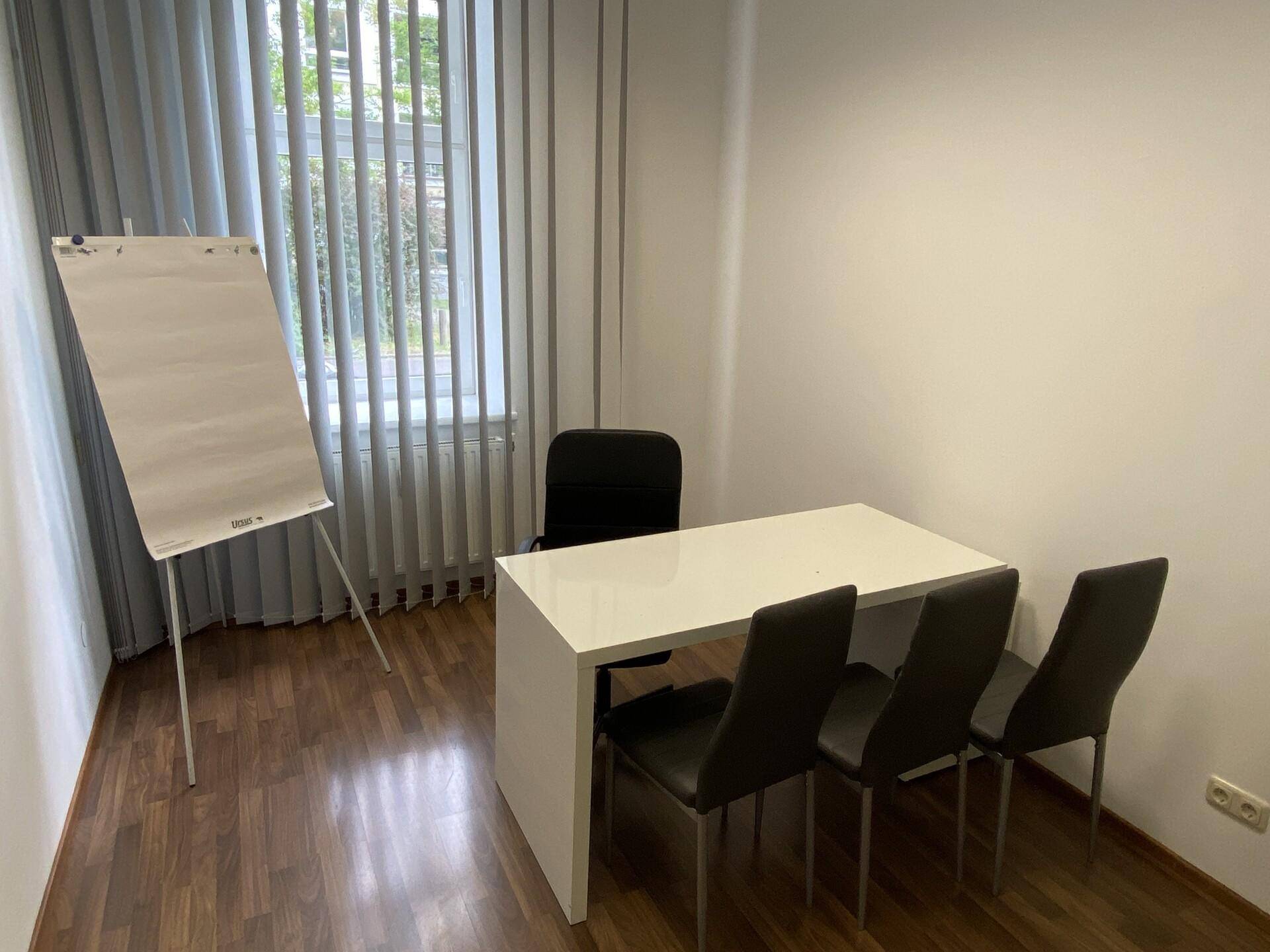 Kleiner Bürobereich mit einem Schreibtisch, drei Stühlen und einem Flipchart neben einem Fenster.