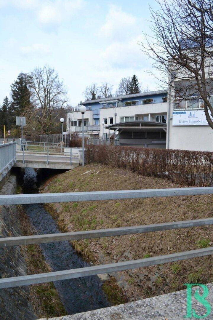 Gebäudeansicht mit angrenzendem Bachlauf und Brücke in ländlicher Umgebung, umgeben von Natur.