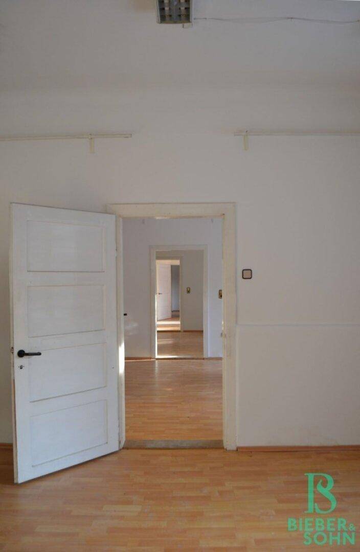 Leerer Raum mit Holzboden und weißen Wänden, offene Türen führen in weitere Zimmer.