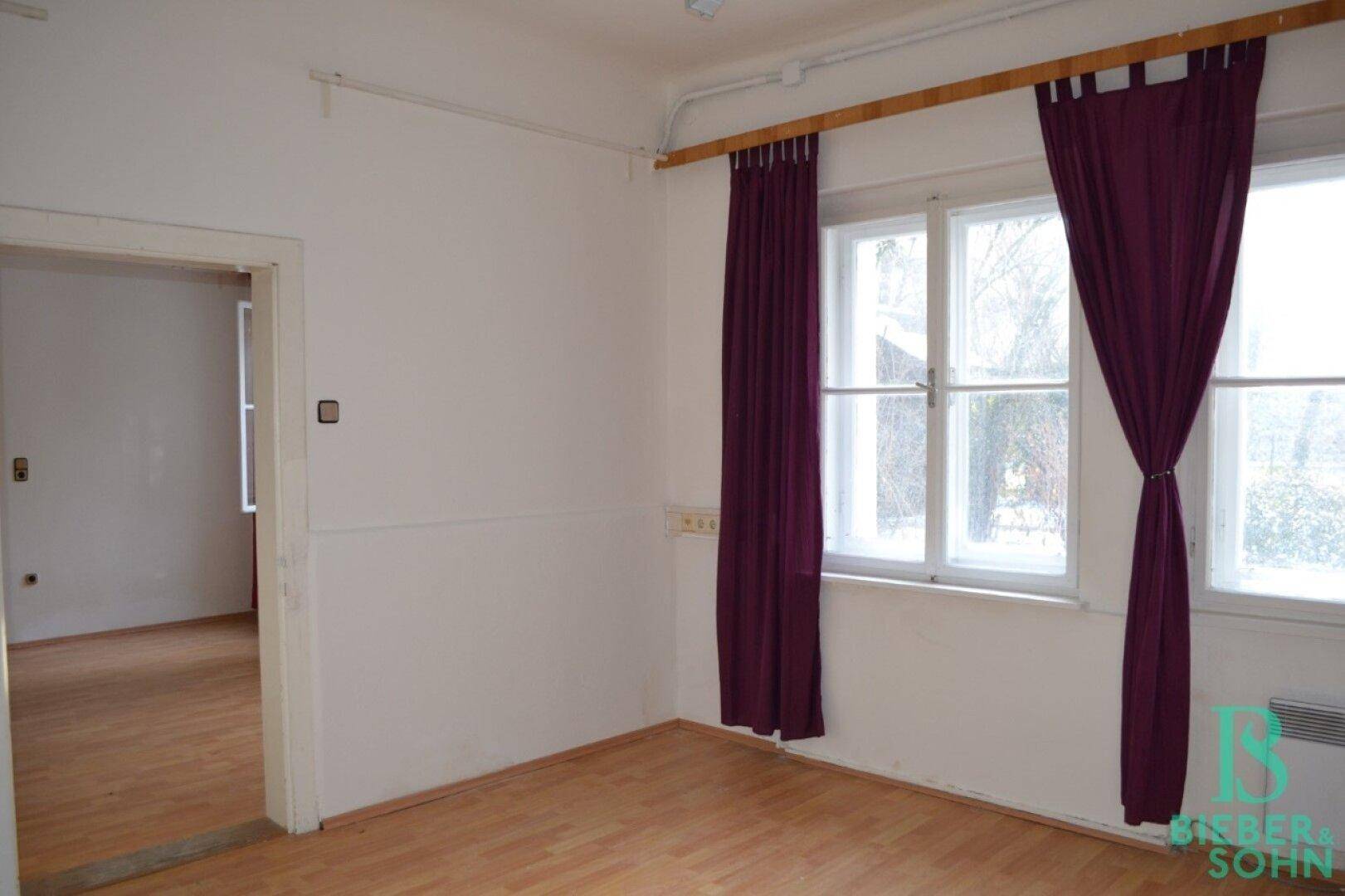 Heller Raum mit Holzboden, weißen Wänden und großen Fenstern mit lila Vorhängen.