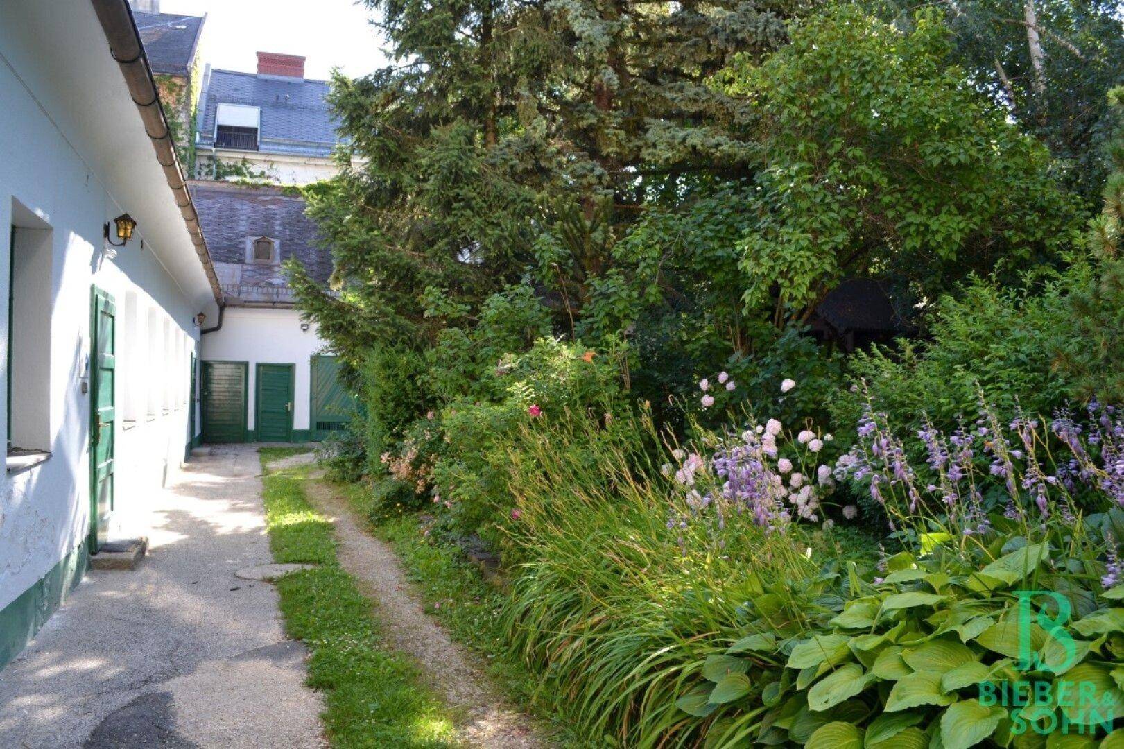 Ein schmaler Gartenweg führt entlang eines Gebäudes, umgeben von üppigem Grün und blühenden Pflanzen.