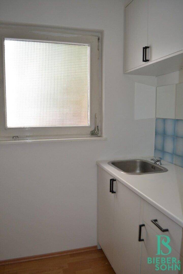 Eine kleine Küche mit weißen Schränken, Spüle und einem Fenster mit Milchglas.