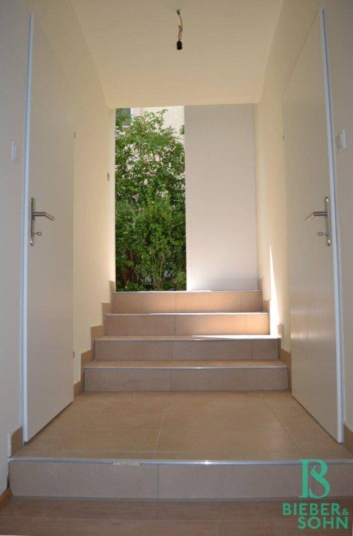 Heller Treppenaufgang mit Fliesenboden, der zu einem grünen Außenbereich führt, flankiert von zwei Türen.