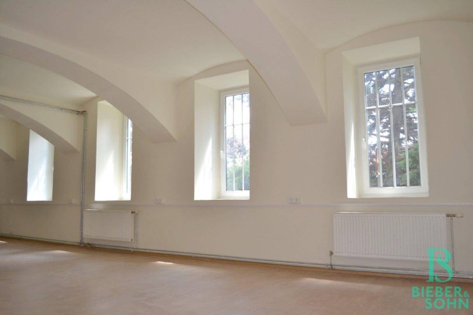 Heller Innenraum mit hohen Fenstern, Gitterstäben und Heizkörpern unter den gewölbten Decken.