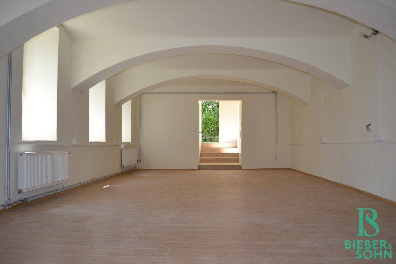 Geräumiger, leerer Innenraum mit gewölbten Decken, hellen Wänden und Blick ins Grüne durch die Tür.