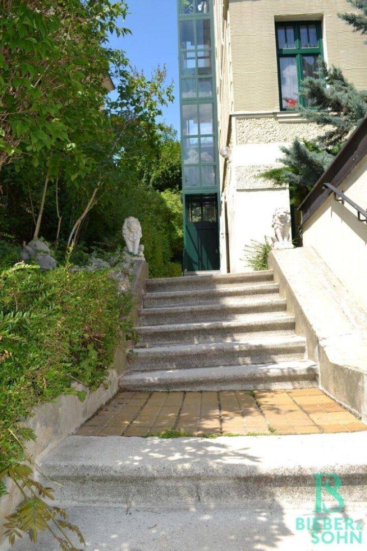 Außentreppe führt zu einem Gebäude mit einem modernen Glasaufzug, umgeben von üppigem Grün.