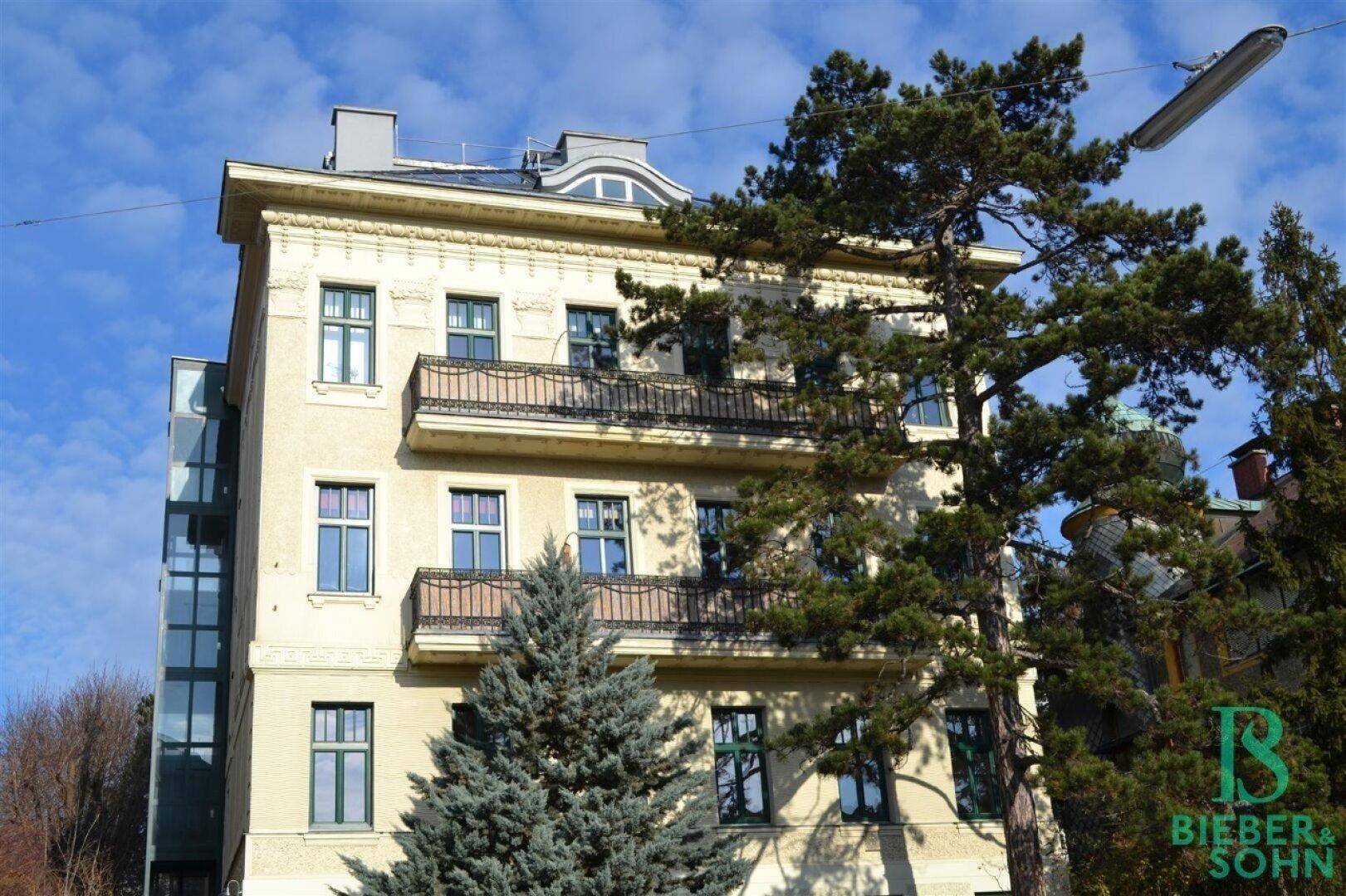 Historisches Gebäude mit Balkonen und einem modernen Glasaufzug an der Seite, umgeben von Bäumen.