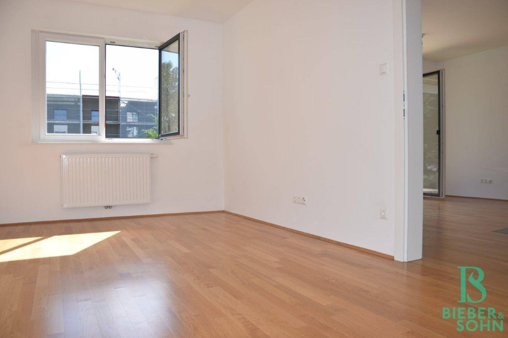 Leerer Raum mit Parkettboden, weiß gestrichenen Wänden und einem Fenster mit Heizkörper darunter.