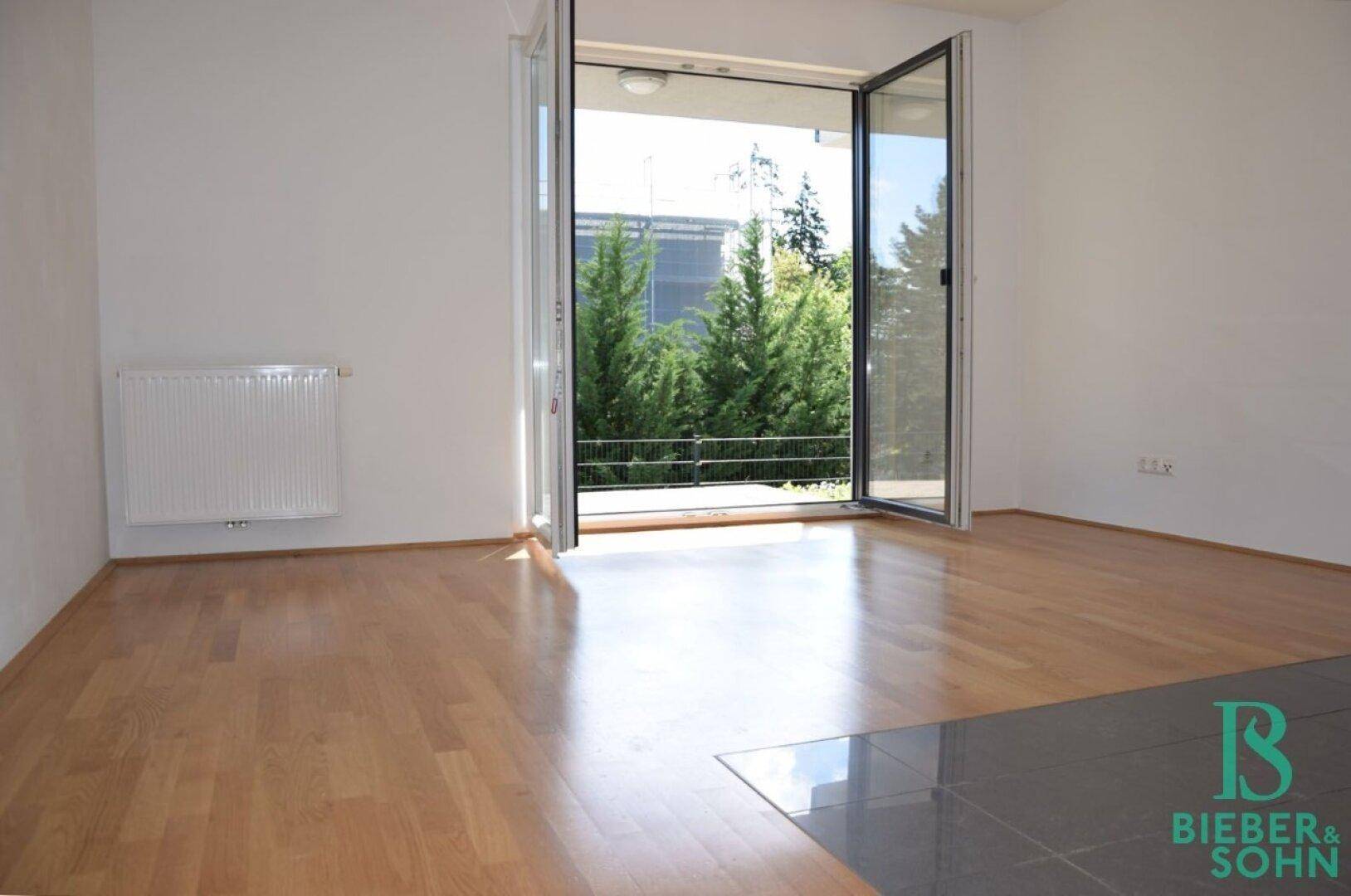 Heller Wohnraum mit Parkettboden, Heizkörper und großen Fenstertüren zum Balkon mit Blick ins Grüne.