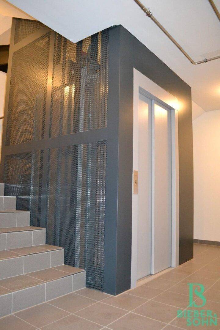 Moderner Treppenaufgang mit Gitterverkleidung und integriertem Aufzug in einem Mehrfamilienhaus.