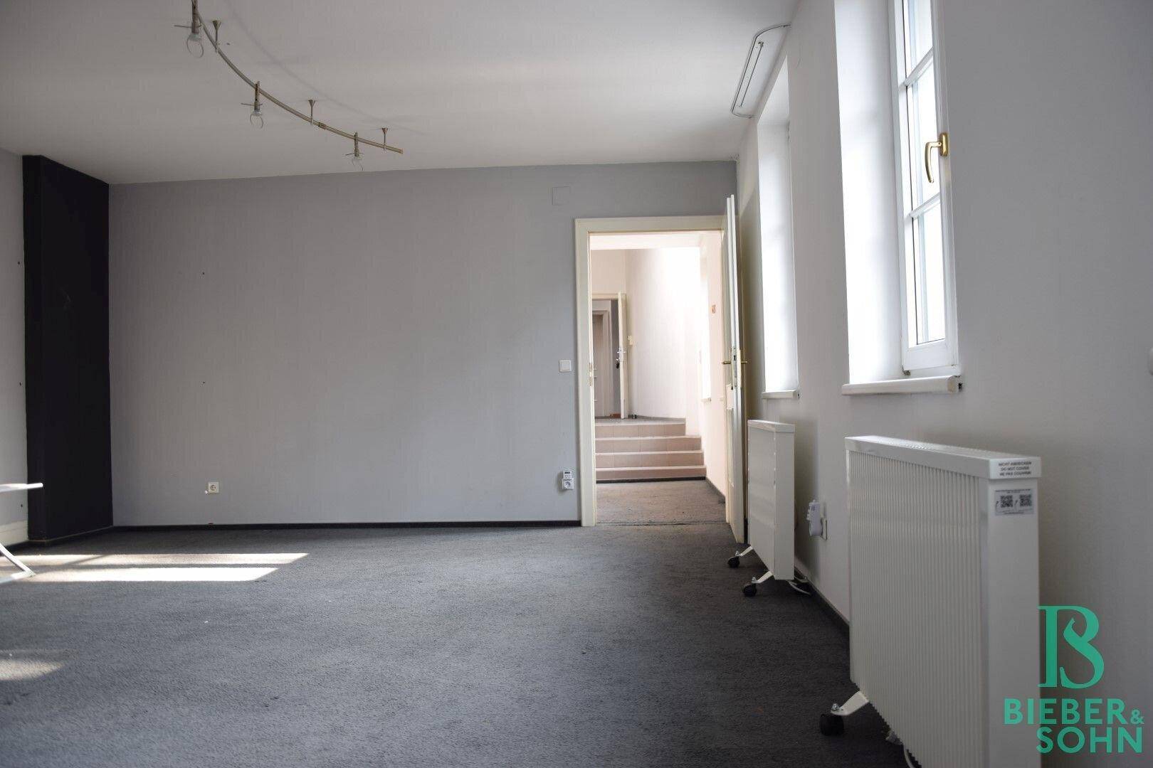 Geräumiger, leerer Raum mit grauem Teppichboden und Fenstern, ausgestattet mit mobilen Heizkörpern.
