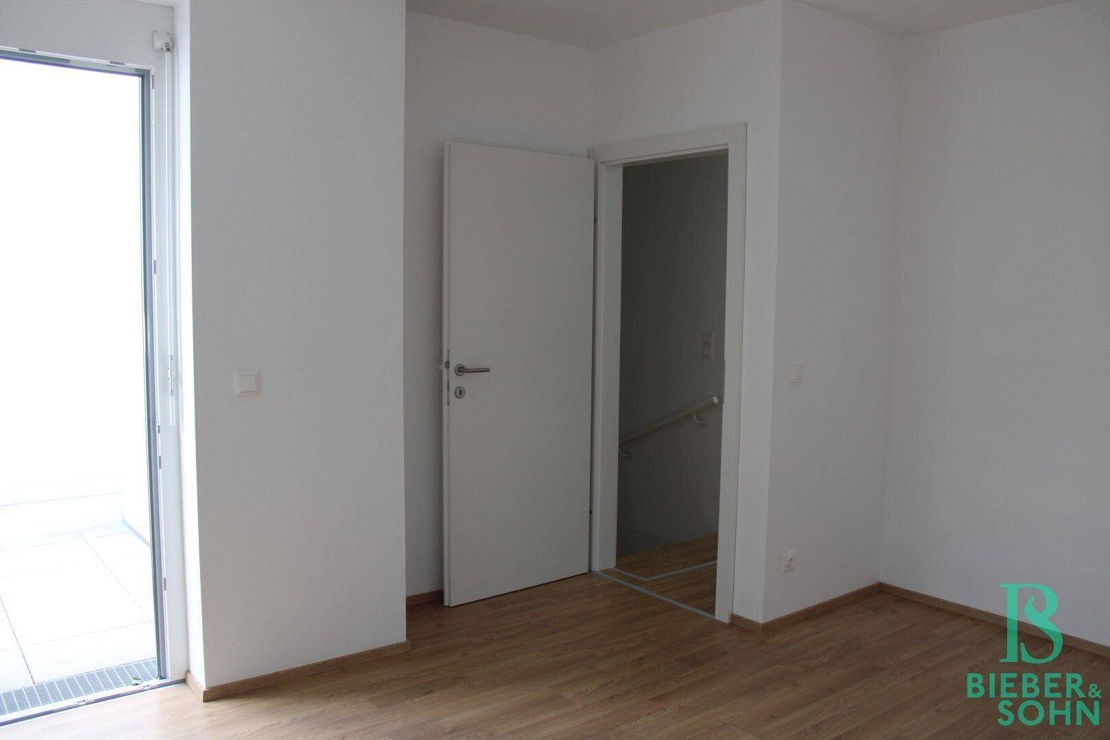 Helles Zimmer mit Holzboden und Zugang zu einem weiteren Raum oder Treppenhaus.