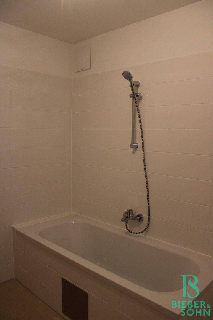 Badezimmer mit Badewanne und Duscharmatur, umgeben von weißen Fliesen.