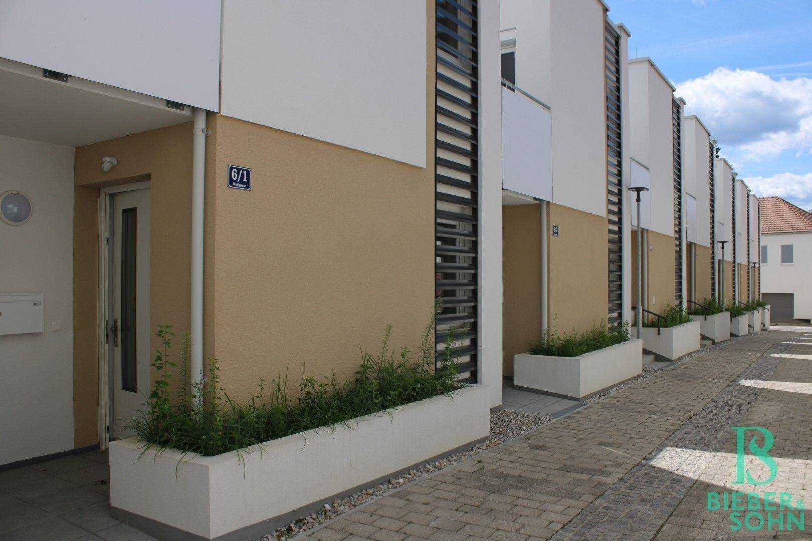 Moderne Reihenhäuser mit beigefarbenen Fassaden und begrünten Pflanzkübeln entlang des Weges.