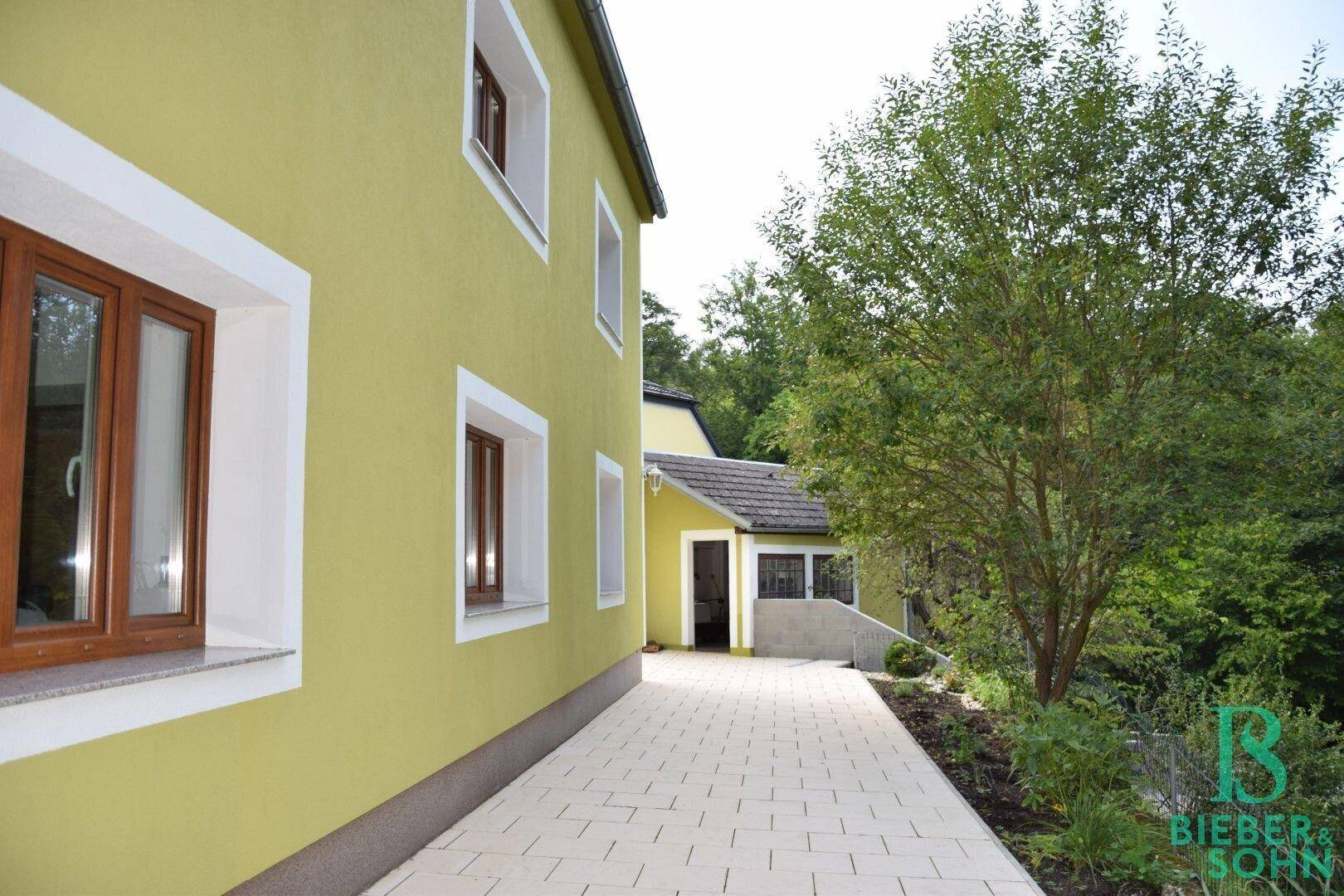 Seitenansicht des Hauses mit grünem Anstrich, gepflastertem Weg und Bäumen.