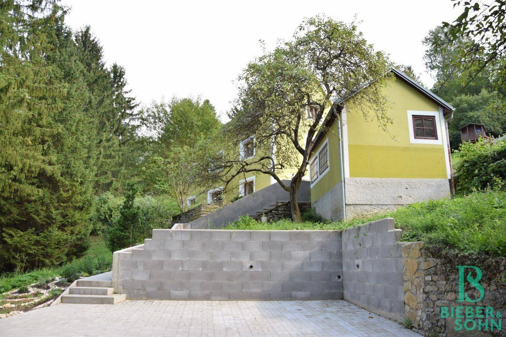 Haus in Hanglage mit Stützmauern, gepflastertem Bereich und umgebender Natur.