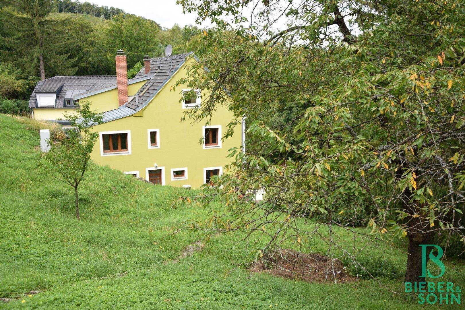 Haus in ländlicher Umgebung, umgeben von Bäumen und grünen Hügeln.