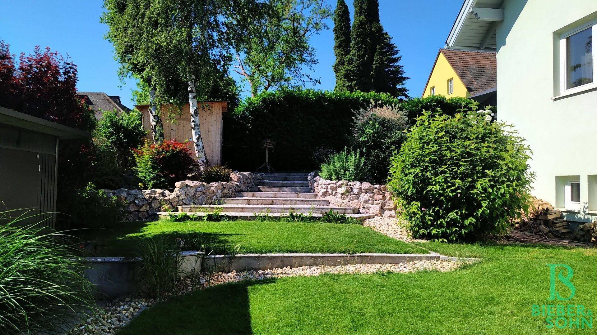 Gestufter Gartenbereich mit Rasenflächen, Steintreppen und üppiger Bepflanzung.