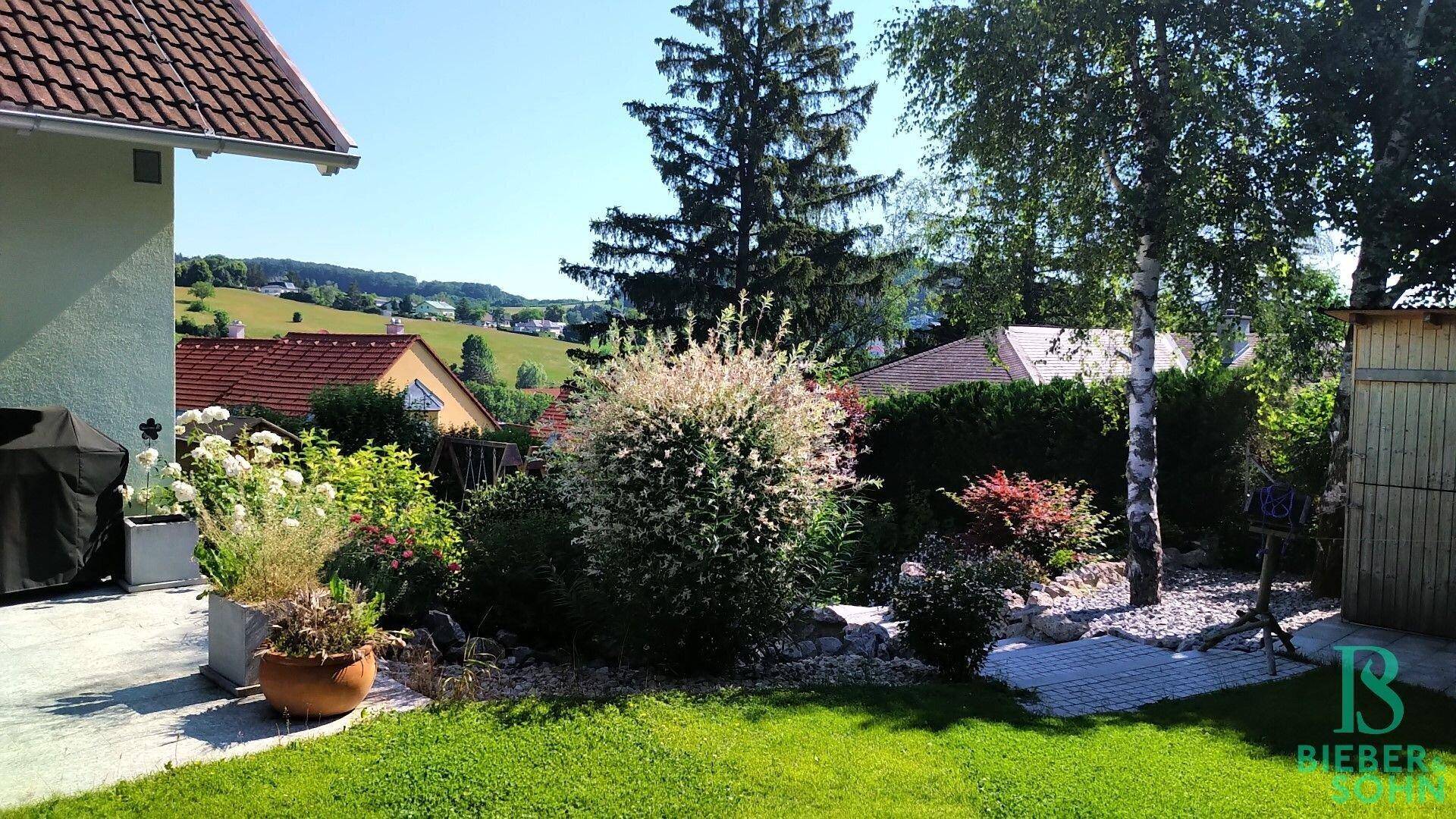 Blühender Gartenbereich mit verschiedenen Pflanzen, Rasen und einem Gartenhaus im Hintergrund.