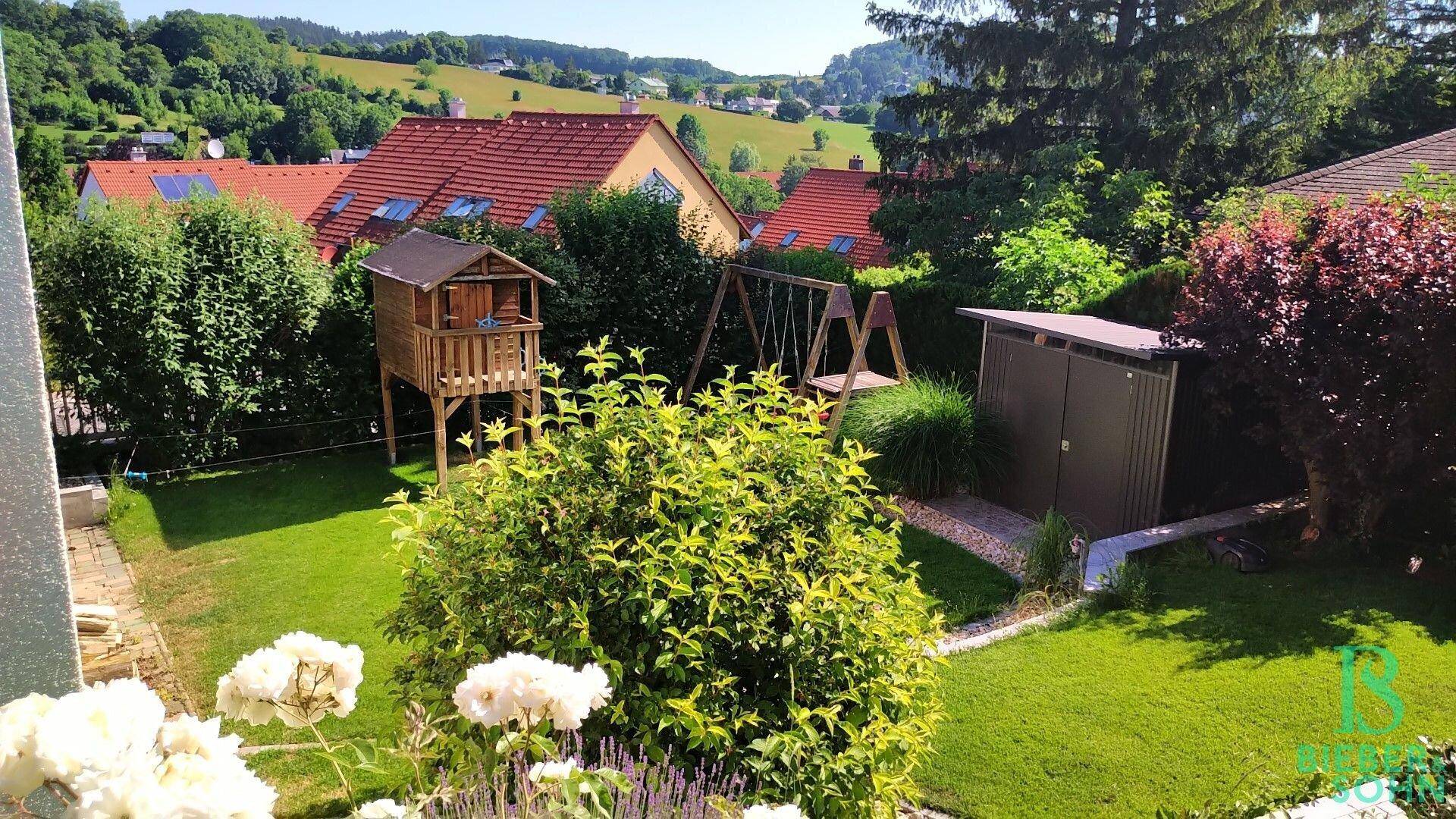 Garten mit Spielplatz, Baumhaus und Schaukel, umgeben von Rasen und Sträuchern.