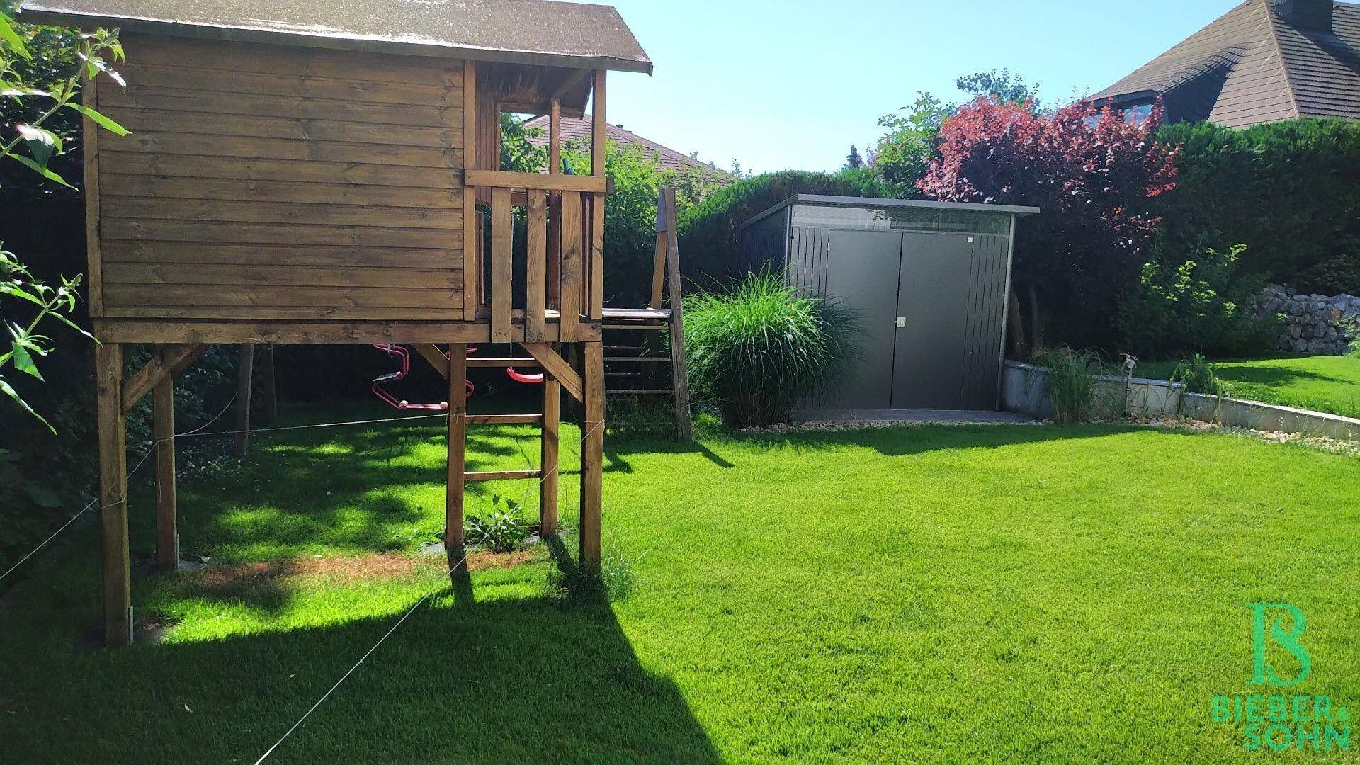 Spielbereich im Garten mit Baumhaus und Rasenfläche, umgeben von Sträuchern.