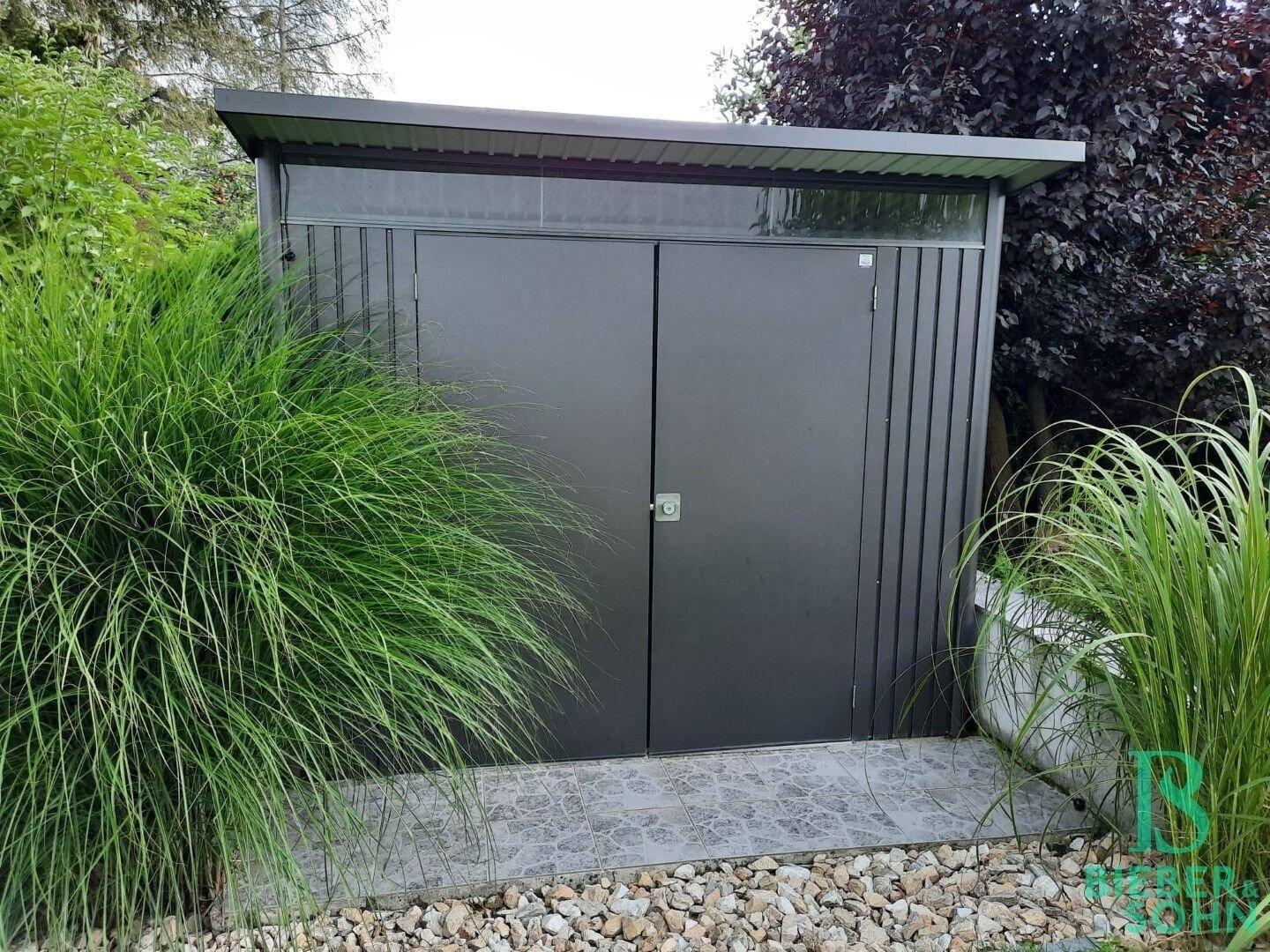 Modernes Gartenhaus aus Metall, umgeben von hohen Gräsern und Sträuchern.