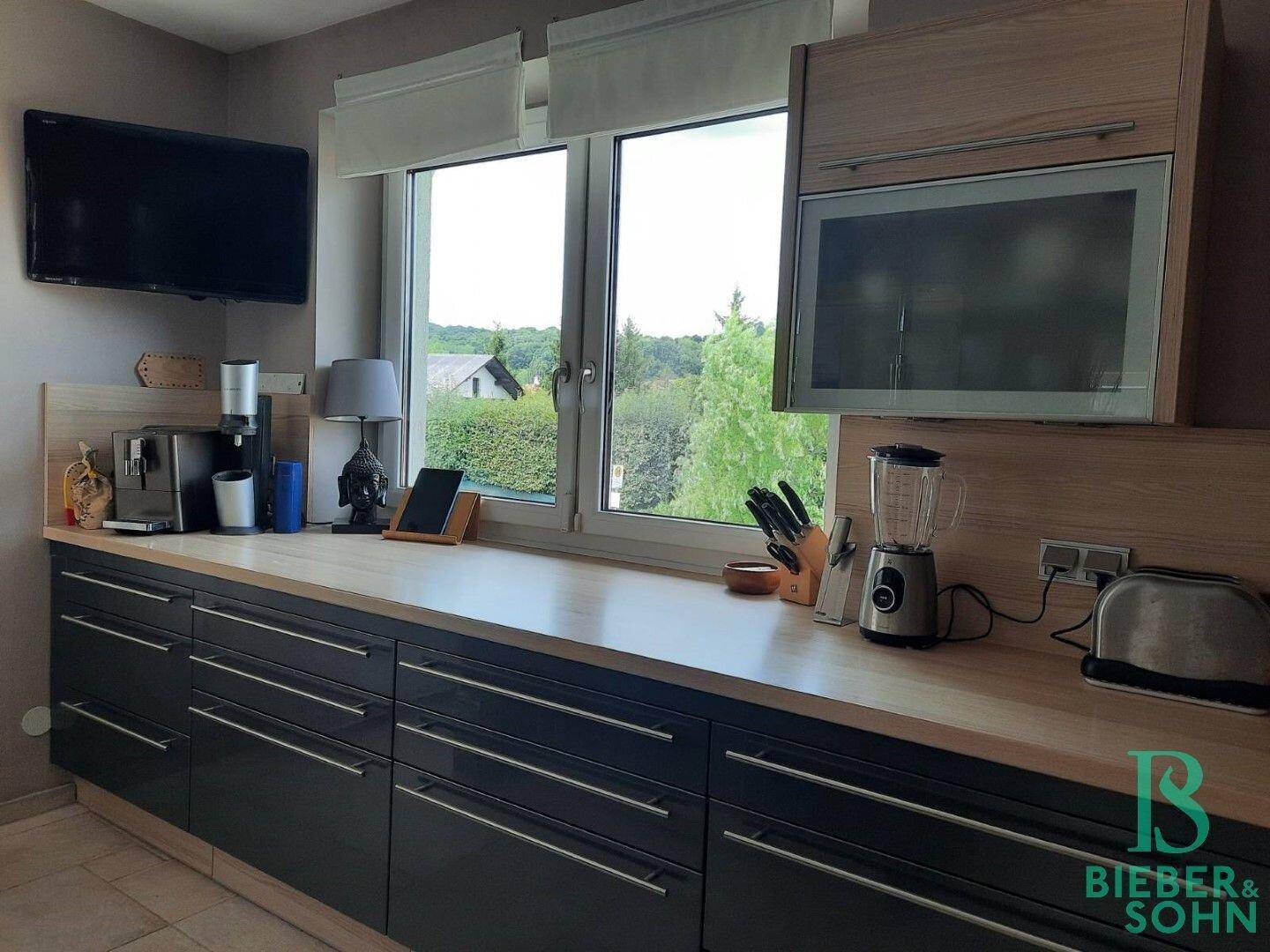 Moderne Einbauküche mit dunklen Fronten, Holz-Arbeitsplatten und Fenster mit Grünblick.