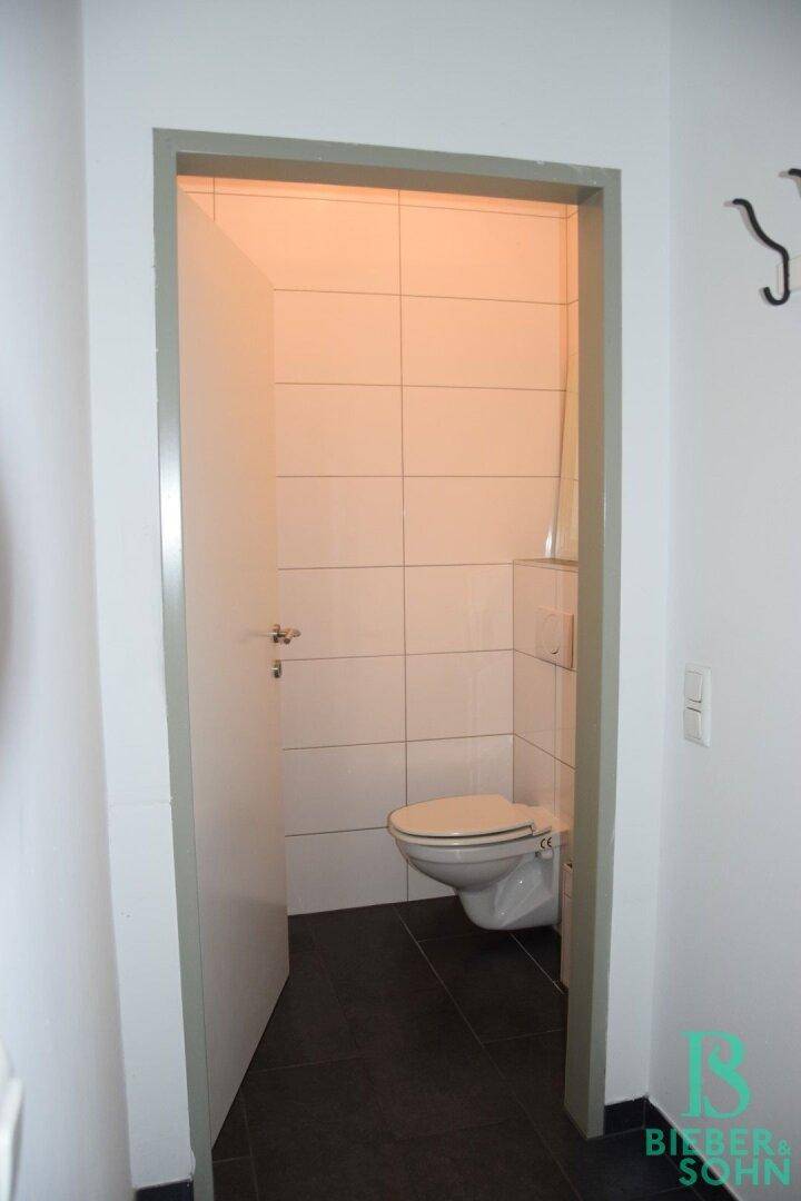Separate Toilette mit wandmontiertem WC und weißen Fliesen, zugänglich über einen Flur.