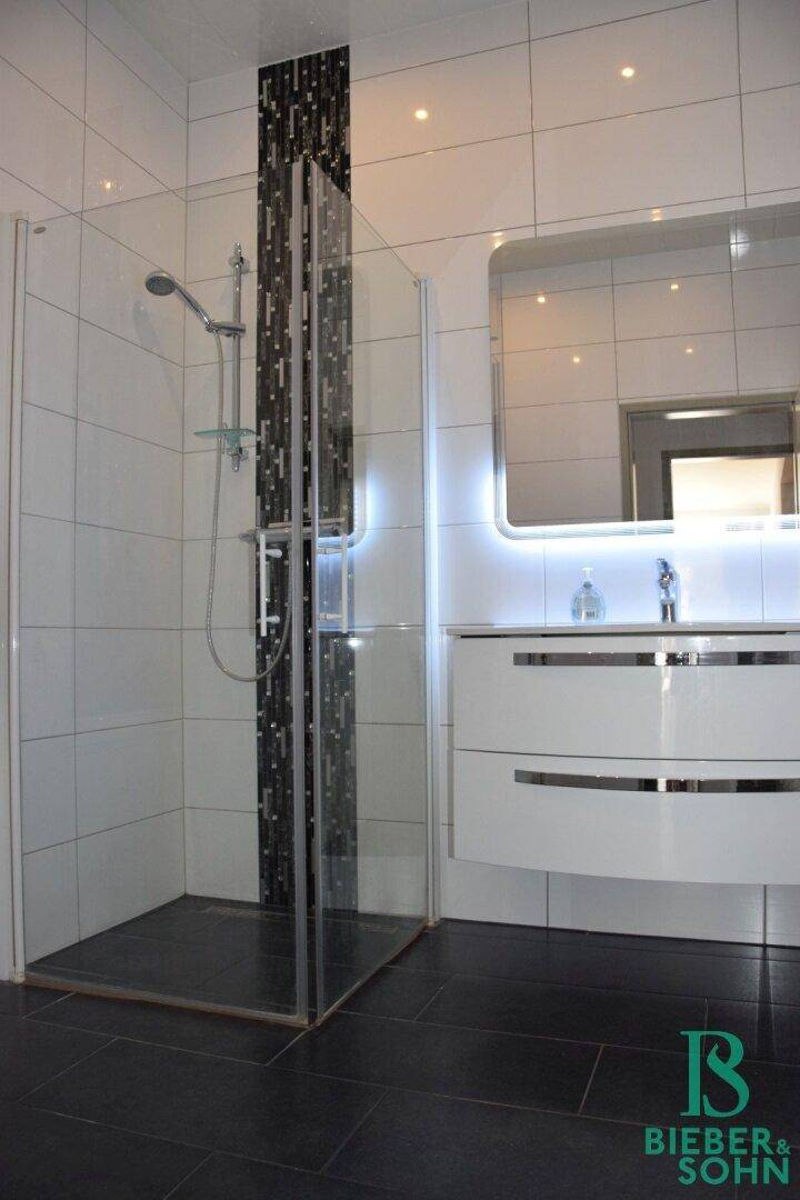 Elegantes Badezimmer mit großer Duschkabine, modernen Fliesen und beleuchtetem Waschtisch.