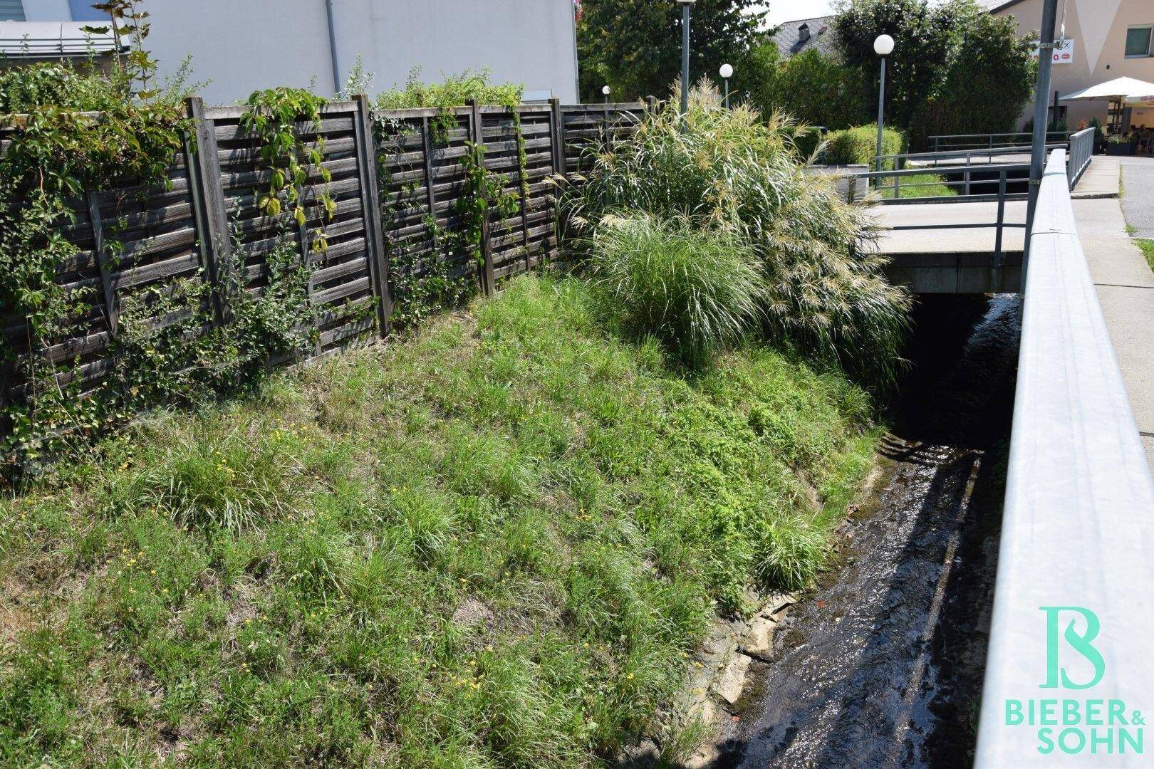 Außenansicht mit einem kleinen Bachlauf, umgeben von Vegetation und einem Holzzaun.