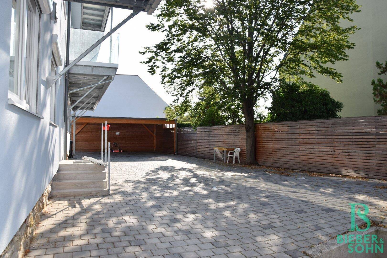 Gepflasterter Außenbereich mit Carport und Holzzaun, umgeben von Bäumen.