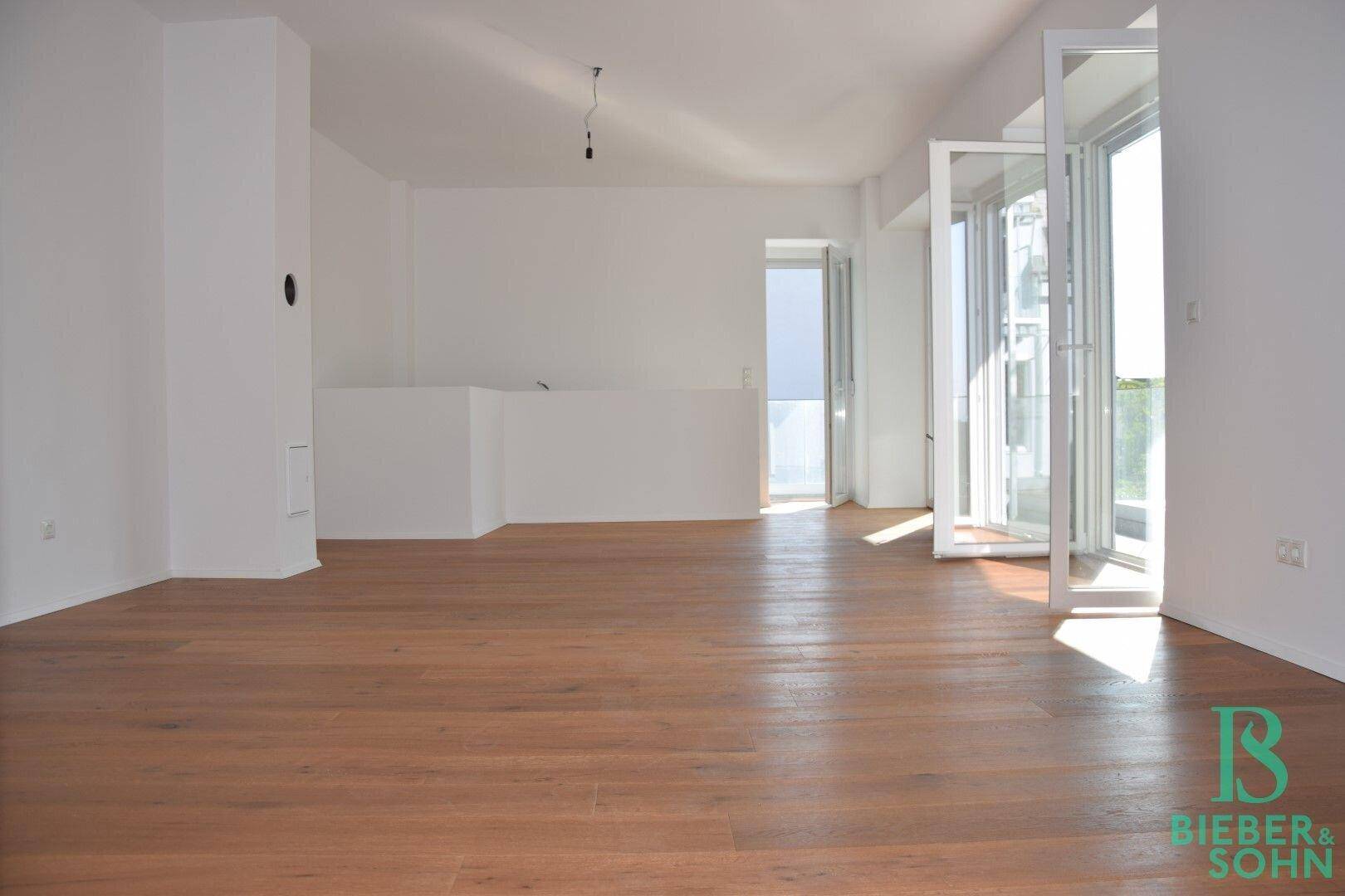 Helles, leeres Wohnzimmer mit Holzboden und großen Fenstern, die viel Tageslicht hereinlassen.