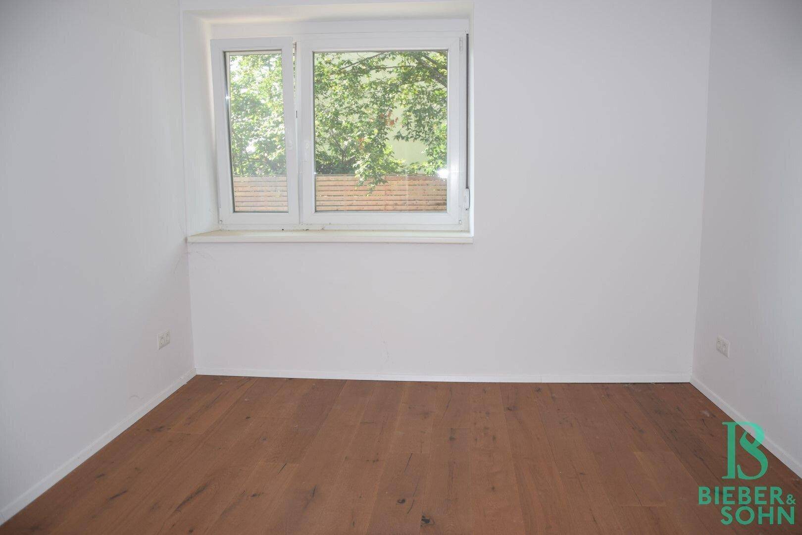 Leeres Schlafzimmer mit Holzboden und Fenster, das viel Tageslicht und Grünblick bietet.