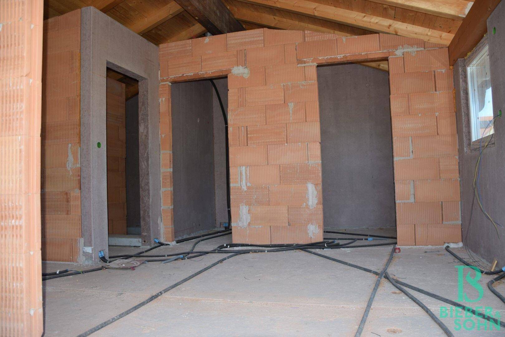 Rohbau-Innenraum mit sichtbaren Ziegelwänden, Holzdecke und vorbereiteten Installationen.