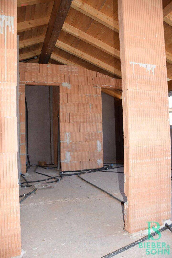 Unfertiger Raum im Dachgeschoss mit sichtbaren Ziegelwänden und Holzdachstuhl.