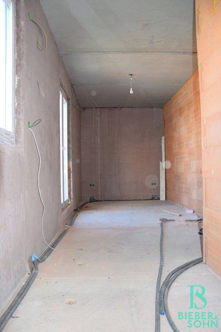 Langer Rohbau-Raum mit Fenstern und sichtbaren Elektroinstallationen.
